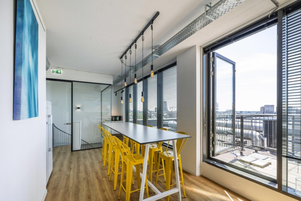 Modern office kitchen with a high table, yellow chairs, hanging lights, and large windows overlooking Elizabeth Cady Stantonplein.